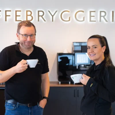 To personer som smiler og holder kaffekopper foran en disk med ordet "kundereferanser" i bakgrunnen.