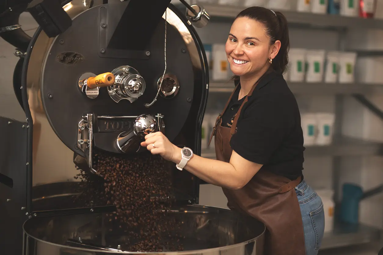 En kvinne i et brunt forkle smiler mens hun betjener en stor kaffebrennemaskin designet for bedrifter i et brenneri.