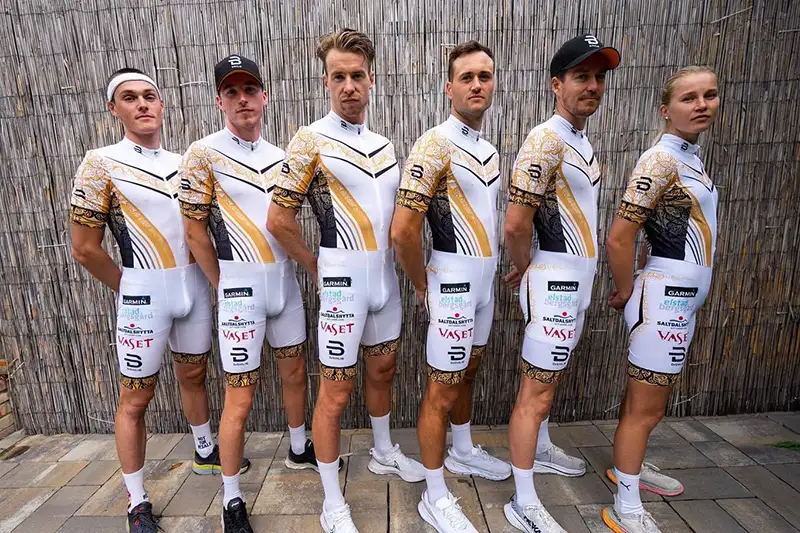 Seks utøvere fra Team Kaffebryggeriet iført matchende sykkeldrakter i hvitt og gull med sponsorlogoer, står selvsikkert foran et bambusgjerde.