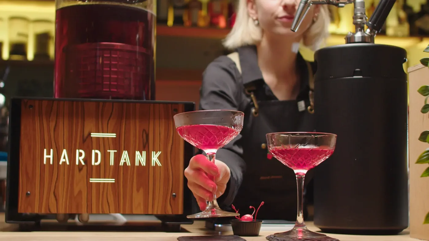 En bartender serverer dyktig rosa cocktailer i kupéglass under et skinnende skilt med teksten «HARDTANK», mens kunder diskuterer de siste kaldbryggetrendene.