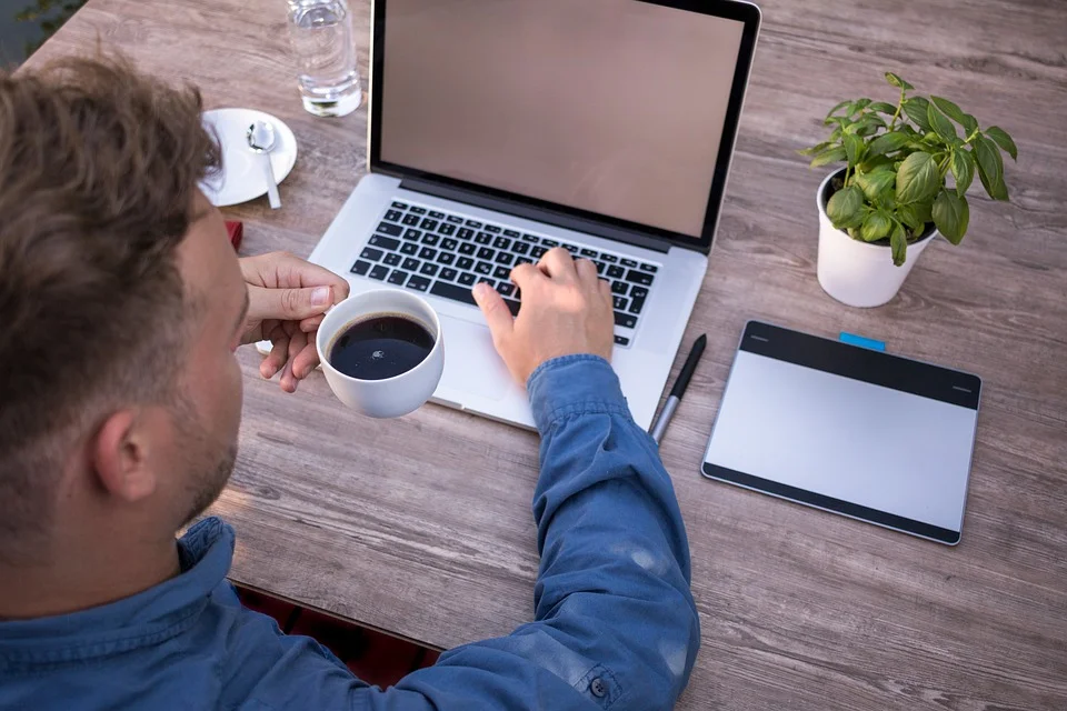 Mann som bruker en bærbar datamaskin på et trebord, holder en kopp kaffe, med et grafisk nettbrett og penn ved siden av datamaskinen, ideell for kaffedrikkere lever lenger.