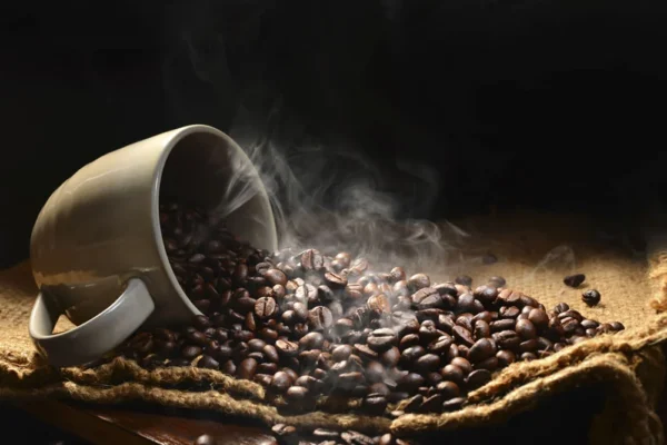 En skråstilt kopp som søler brente kaffebønner og avgir damp på en burlap-overflate med mørk bakgrunn, som antyder at kaffedrikkere lever lenger.