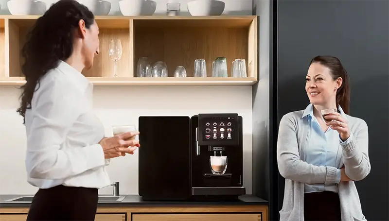 To kvinner som smiler og snakker, holder kopper nær en Franke A300 kaffemaskin på et kjøkken med åpne hyller.