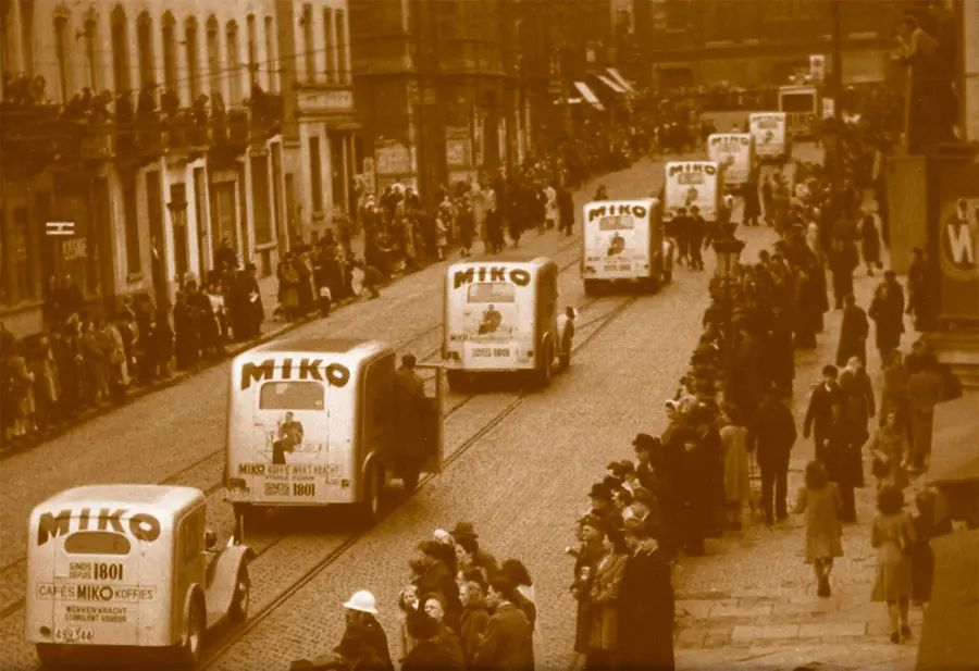 Historisk sepia-tonet bilde av en gate med folk i kø og ser på en parade med vintage miko-isbiler, organisert av Om Kaffebryggeriet.