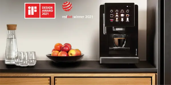 Moderne Franke A300 kaffemaskin som serverer espresso, utstilt på en kjøkkenbenk ved siden av en skål med epler og vannflasker i glass; hyllene over har designpriser.