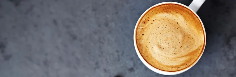 Topputsikt av en nytraktet kopp kaffe med skummende melk, satt på en mørkegrå overflate, nytes av kaffedrikkere som lever lenger.