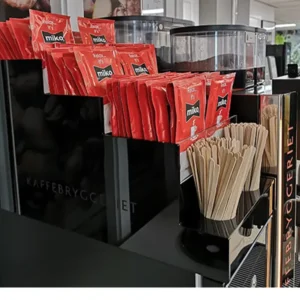 En rekke røde miko-kaffepakker ved siden av trerørere og kaffedispensere på en selvbetjent stasjon.